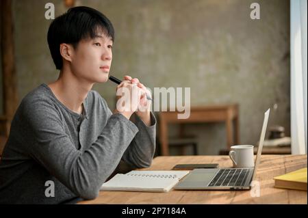 Jeune homme asiatique attentionné et concentré, vêt de vêtements décontractés, regardant par la fenêtre, pensant et planifiant quelque chose, rêvant de la journée tout en travaillant à distance Banque D'Images