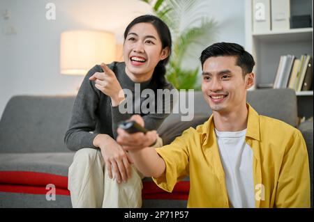 Un jeune couple asiatique joyeux et joyeux rit tout en regardant la télévision dans le salon, passant du temps ensemble à la maison. petit ami an Banque D'Images