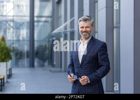 Portrait d'un homme d'affaires heureux et souriant aux cheveux gris, homme en costume d'affaires debout à l'extérieur du bâtiment de bureau souriant et regardant l'appareil photo, investisseur de boss mûr satisfait tenant des lunettes. Banque D'Images