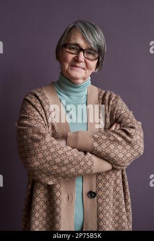 Portrait vertical à la taille de la femme âgée portant un gilet et des lunettes regardant l'appareil photo tout en se tenant debout avec les bras croisés Banque D'Images
