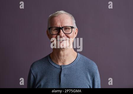 Portrait minimal de l'homme âgé portant des lunettes et souriant à l'appareil photo sur fond violet, espace de copie Banque D'Images
