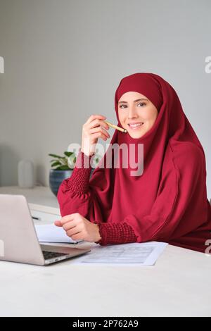 Portrait de la jeune femme musulmane dans le hijab utilisant un ordinateur portable à la maison. Banque D'Images