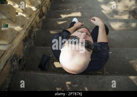 Jeune homme beau habillé décontracté assis sur les escaliers dans un parc. Photo en hauteur en regardant vers le bas sur un gars cool bald dans des lunettes de soleil se détendre à l'extérieur Banque D'Images