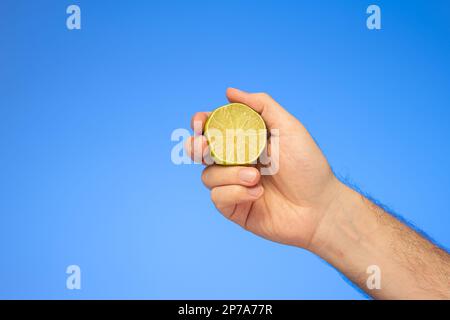 Vert frais moitié citron vert tenu à la main par le mâle caucasien main gros plan isolé sur fond bleu. Banque D'Images