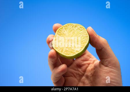 Vert frais moitié citron vert tenu à la main par le mâle caucasien main gros plan isolé sur fond bleu. Banque D'Images