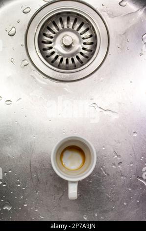 Petite tasse de café sale en porcelaine blanche dans un évier de cuisine en métal à côté du trou d'évacuation vue du dessus. Banque D'Images