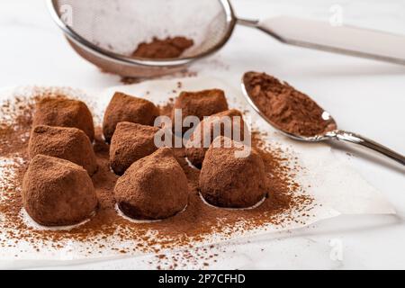 Truffes au chocolat saupoudrées de poudre de cacao en gros plan. Délicieuses boules de bonbons au chocolat noir, tamis en maille et cuillère sur une surface en marbre. Macro. Banque D'Images