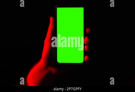 Téléphone portable dans la main jeune homme. Utilisez un smartphone avec écran vert sur un éclairage au néon bleu rouge et un arrière-plan sombre. Affichage de la touche noire du téléphone sur un homme Banque D'Images