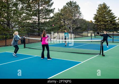 Au début du printemps, quatre joueuses de pickleball vollent une balle rose près du filet sur un terrain bleu et vert. Banque D'Images