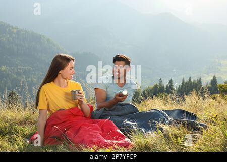 Couple heureux avec des boissons dans des sacs de couchage entouré par la belle nature Banque D'Images