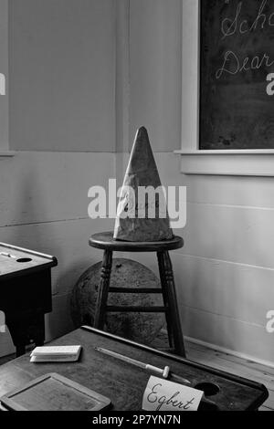 Un chapeau de dounce se trouve sur un tabouret dans le coin d'une école locale de 1750 pièces, avec plusieurs bureaux pour étudiants, un bureau pour enseignants et un blackboar Banque D'Images