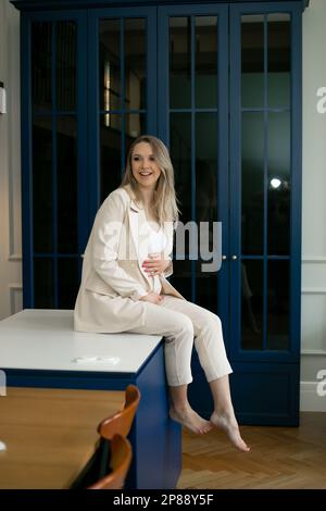 Une femme d'affaires charmante et heureuse est assise à la maison sur fond de portes bleues et épouse doucement son estomac. Banque D'Images