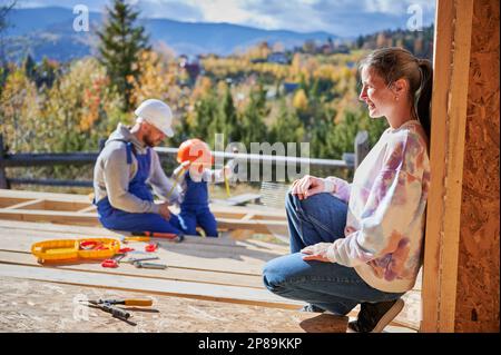 Père, mère et fils, maison en bois. Un jeune garçon aidant son papa, alors que la femme à la recherche d'eux sur le chantier de construction. Les gars portant un casque et une combinaison bleue. Menuiserie, concept de famille. Banque D'Images