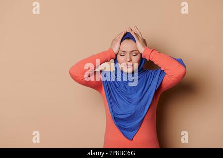 Femme musulmane dans le hijab, frottant les temples, traitant les maux de tête de tension, souffre de migaine, grimacing dans la douleur, toile de fond beige Banque D'Images