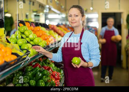 Une vendeuse d'âge moyen met des pommes dans une étaie alimentaire dans une épicerie Banque D'Images