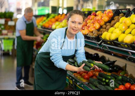 Une vendeuse d'âge moyen mettant des concombres dans une étaie alimentaire à la greengrocery Banque D'Images