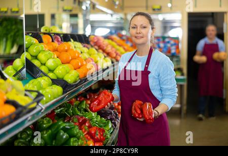 Femme professionnelle adulte vendeur en tablier mettant les poivrons rouges et verts sur les étals dans hypermarché Banque D'Images