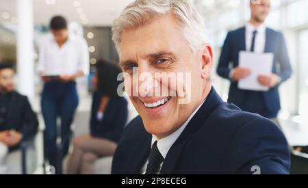 Pouvez-vous dire à quel point je suis heureux ? Portrait court d'un beau homme d'affaires mûr assis et souriant tandis que ses collègues travaillent derrière lui dans le bureau. Banque D'Images