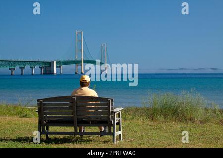Un long pont suspendu avec un homme assis sur un banc de parc en premier plan. Banque D'Images