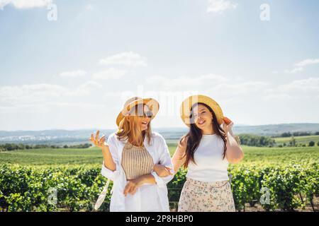 Deux jeunes femmes heureuses embrassent et marchent à travers le vignoble. Les amies blanches en vêtements décontractés et chapeaux de paille regardent le paysage. Amitié a Banque D'Images