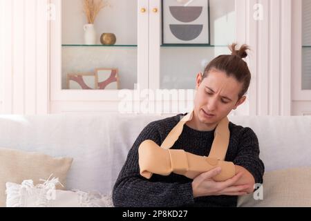 Jeune homme malheureux dans la douleur avec une main cassée portant une élingue de bras assis sur un canapé dans le salon à la maison Banque D'Images