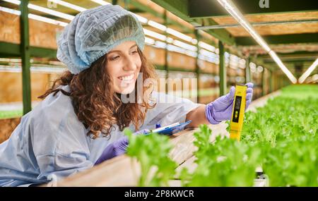 Jardinière féminine joyeuse dans un capuchon jetable tenant le presse-papiers et souriant tout en mesurant la valeur de pH du sol avec un appareil électronique numérique. Femme scientifique utilisant un pH-mètre en serre Banque D'Images