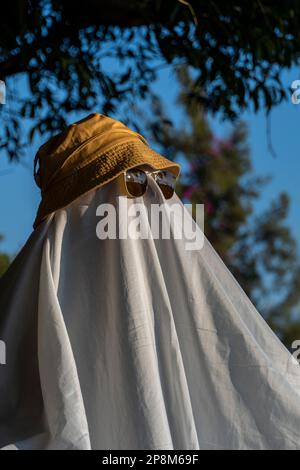 Une personne portant un chapeau et des lunettes de soleil à rebord noir sur un tissu blanc ressemblant à un fantôme Banque D'Images