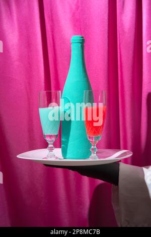 Un serveur en gants noirs tient un plateau blanc avec un verre de vin rouge et turquoise et une bouteille de champagne. Arrière-plan des rideaux magenta. Banque D'Images