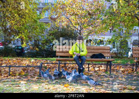 un garçon dans une veste jaune avec des lunettes est assis sur un banc et nourrit une goubei Banque D'Images