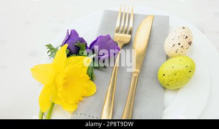 Table de Pâques, œufs de Pâques, décoration de fleurs fraîches du printemps, serviette et couverts dorés sur fond blanc, au-dessus. Banque D'Images