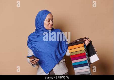 Femme musulmane arabe en hijab bleu, décorateur maison posant avec un smartphone et des échantillons de tissus d'ameublement sur fond beige Banque D'Images