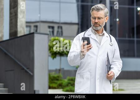 Un médecin de sexe masculin plus âgé aux cheveux gris est debout à l'extérieur de la clinique dans un uniforme et tient une chemise. Utilise un téléphone mobile, lit avec inquiétude les messages, appelle un patient, chat. Banque D'Images