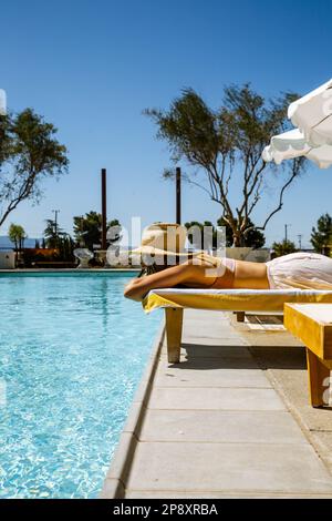Une belle femme en vacances se trouve sur une chaise longue à côté d'une piscine bleue portant un chapeau de soleil. Elle repose la tête dans ses bras croisés en regardant dehors. Banque D'Images
