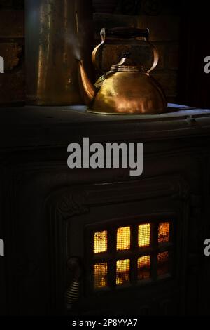 Une bouilloire en cuivre brillante avec de la vapeur qui s'échappe, s'est assise sur un vieux poêle à bois en fonte avec une flamme jaune et orange, dans un éclairage doux et sombre Banque D'Images