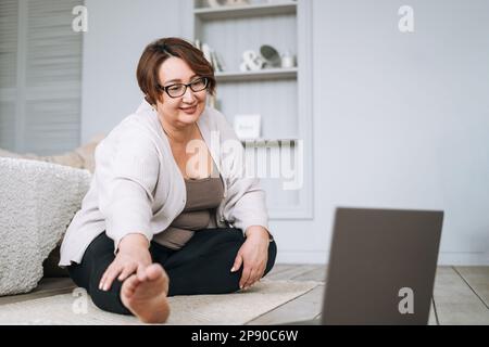 Adulte charmante femme brune plus taille corps pratique positive yoga avec ordinateur portable à la maison Banque D'Images