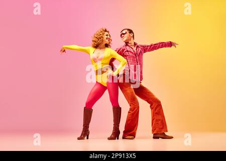 Danse disco. Elégant et expressif couple enthousiaste de danseurs professionnels dans des vêtements de style rétro dansant sur fond rose-jaune. Banque D'Images