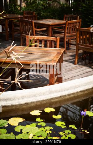 Terrasse extérieure avec tables et chaises en bois anciennes et miteuses au soleil. Étang aux nénuphars sur une terrasse romantique du restaurant Banque D'Images