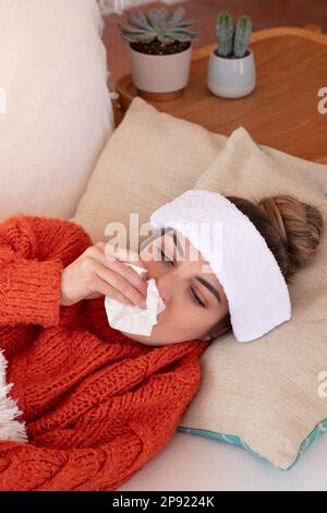 Femme malwell avec fièvre froide soufflant nez avec papier mouchoir tout en se reposant avec une serviette humide sur la tête Banque D'Images