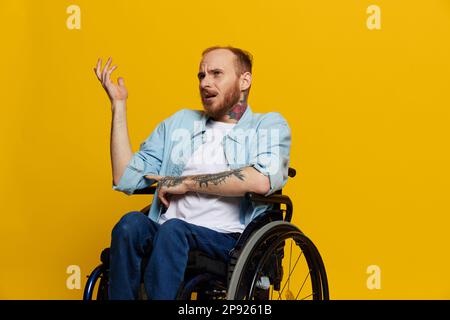 Un homme dans un fauteuil roulant problèmes avec le système musculo-squelettique regarde la caméra n'est pas satisfait, avec des tatouages sur ses bras assis sur un studio jaune Banque D'Images