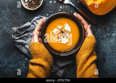 Les mains des femmes sont en tricot jaune et tiennent un bol avec une soupe à la crème de potiron sur fond de pierre sombre avec une cuillère décorée de citrouille fraîche coupée Banque D'Images