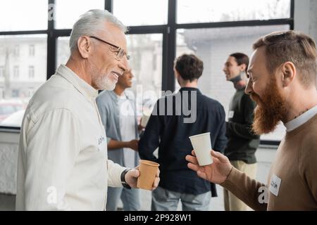 Hommes souriants avec des tasses en papier parlant pendant la réunion des alcooliques dans le centre de récupération, image de stock Banque D'Images