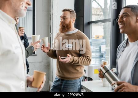 Homme barbu avec une tasse en papier et un smartphone parlant aux gens pendant la réunion des alcooliques dans le centre de récupération, image de stock Banque D'Images