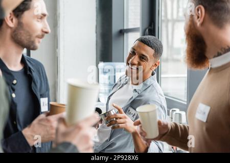 Homme afro-américain joyeux tenant des thermos et parlant à des gens pendant la réunion des alcooliques dans le centre de réadaptation, image de stock Banque D'Images