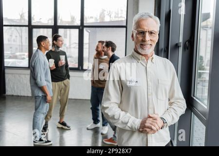 Homme d'âge moyen souriant regardant la caméra près d'un groupe flou pendant la réunion des alcooliques dans le centre de réadaptation, image de stock Banque D'Images