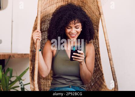 Femme noire, téléphone et sourire pour la communication, les médias sociaux et la diffusion en ligne avec le wi-fi dans un fauteuil en bois rustique balançoire. Femme avec un sourire Banque D'Images