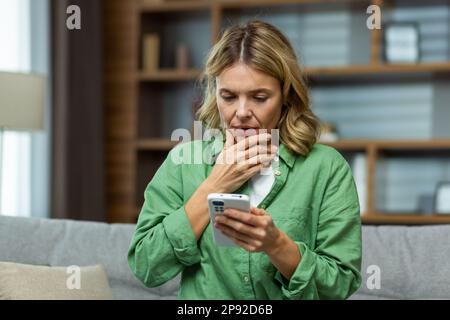 Gros plan. Femme âgée inquiète mère assis sur un canapé à la maison et tenant le téléphone. S'inquiète des enfants, écrit et envoie des messages, des appels, des recherches, des attentes à la maison. Banque D'Images