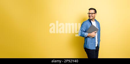 Homme souriant tenant un ordinateur portable et regardant par-dessus l'épaule. Un homme charmant et gai debout sur fond jaune. Il porte une tenue décontractée Banque D'Images