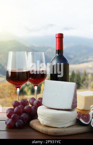 Différents types de fromages délicieux, en-cas et vin sur une table en bois contre le paysage de montagne Banque D'Images