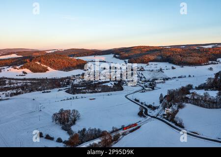 Allemagne, Thuringe, Bechstedt, train régional 60, train 29889 approche de l'arrêt Bechstedt-Trippstein dans les derniers rayons du soleil, maison (ancien bâtiment d'accueil), paysage, village, champs, forêts, neige, vue d'ensemble, lumière du soir, photo aérienne Banque D'Images