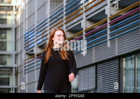 Une jeune femme aux cheveux rougeâtres se promène volontiers à travers un complexe avec des maisons de style architectural moderne Banque D'Images